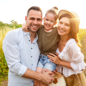 A smiling man, woman, and young girl pose together outdoors in a sunny field. The girl is held between the adults, all looking happy. The woman wears a straw hat and holds a woven bag.