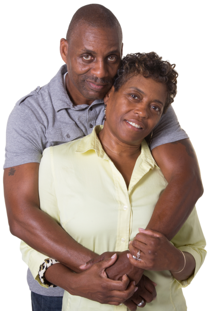 A man in a gray shirt hugs a smiling woman in a light yellow blouse from behind. Both look at the camera, and the woman—who lives joyfully despite her hearing loss—holds his hands gently. The background is plain white.